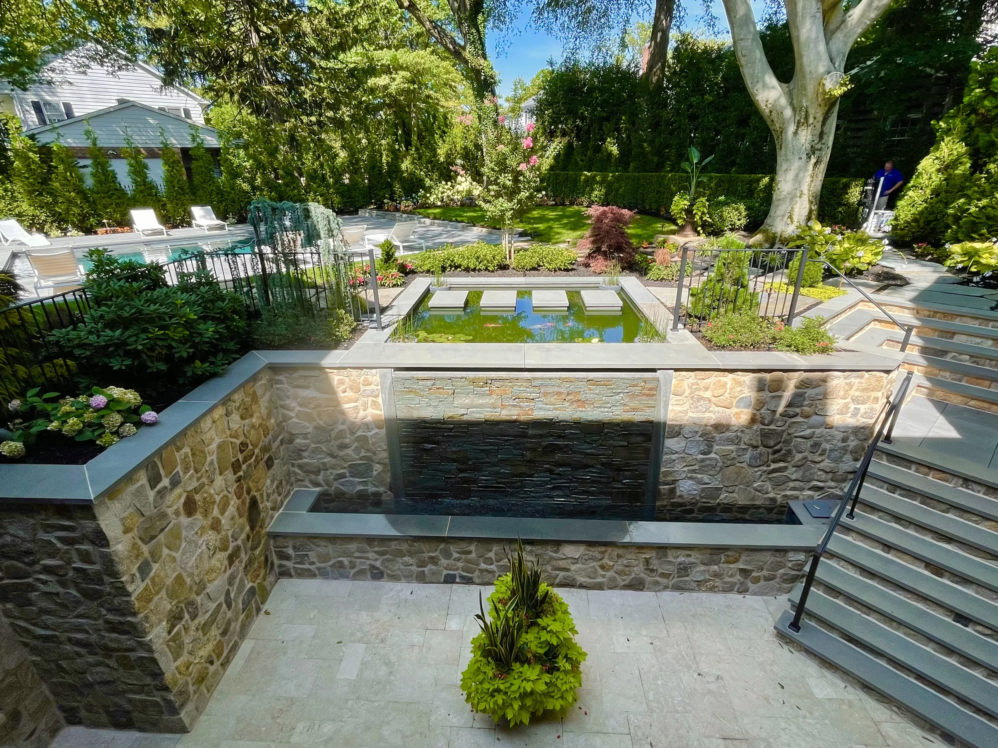 Stone terrace with a water wall, planting beds, and steps beside the pool.