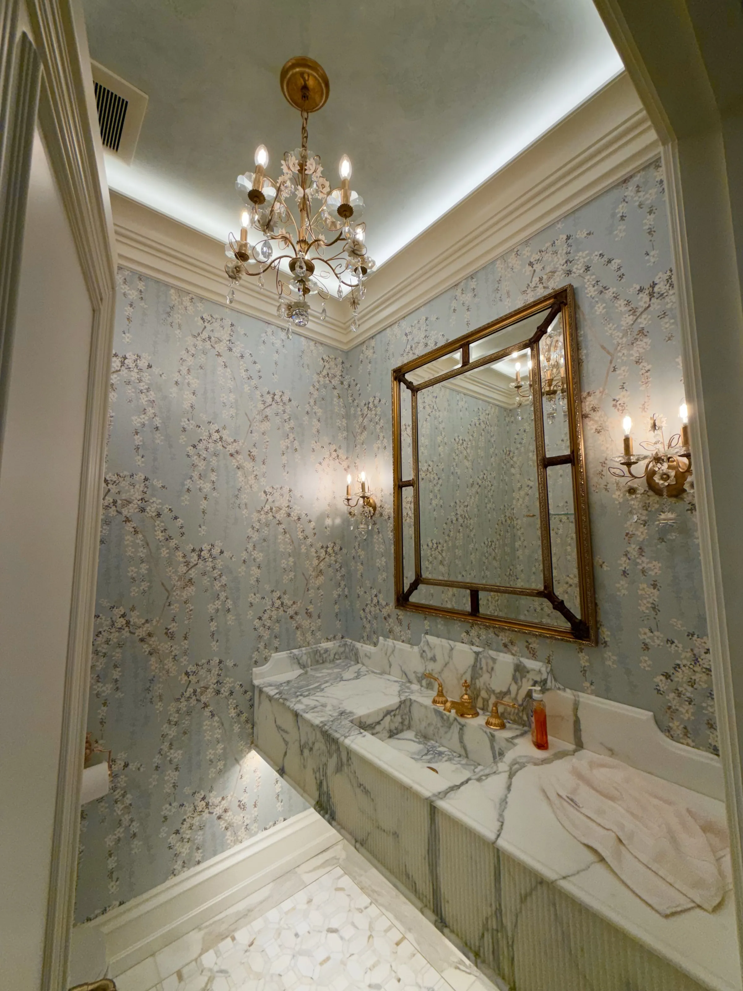 Powder room with a marble vanity, patterned walls, and brass sconces.